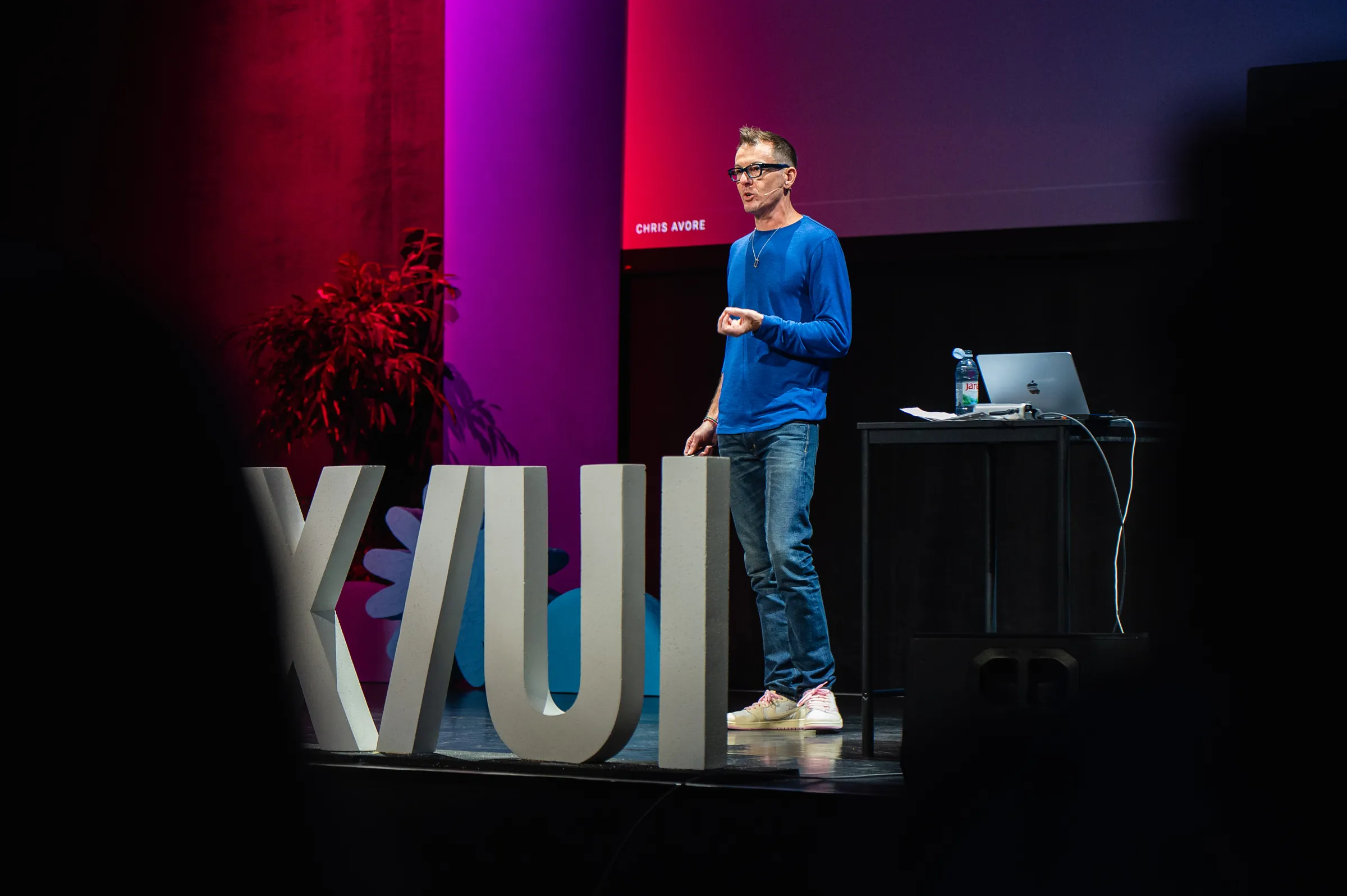 Chris Avore speaking on a stage lit in magenta and blue, in a blue sweater and jeans, standing behind oversized concrete letters, with a laptop on a table to his right.